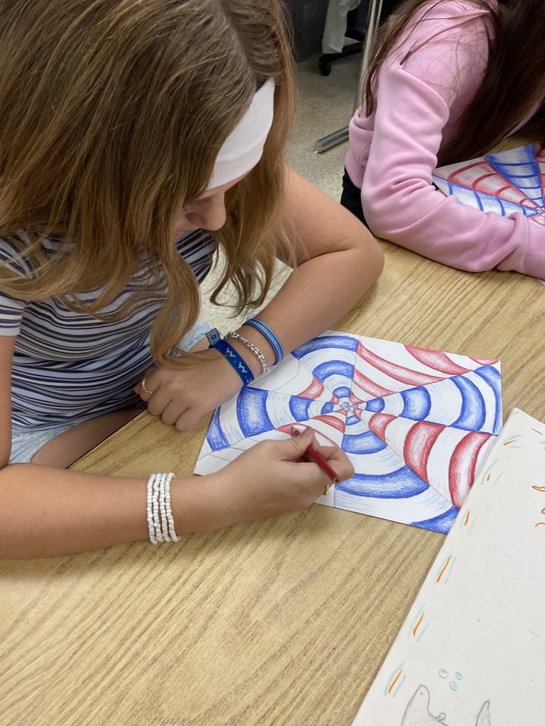 Student coloring a geometric optical illusion drawing with red and blue colored pencils at a classroom table.