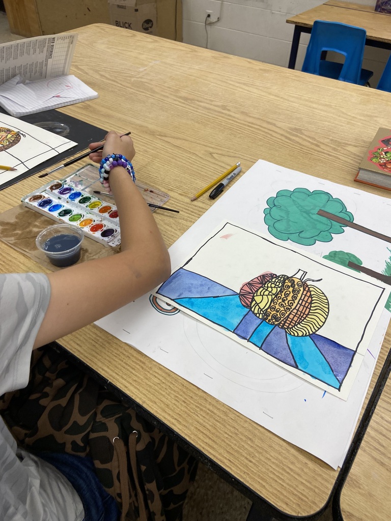 Student painting a colorful art project at a classroom table with watercolor paints and drawing supplies.