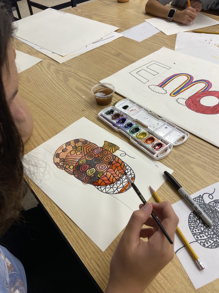 Student painting a patterned pumpkin artwork at a classroom table with watercolor paints, pencils, and other student art projects.