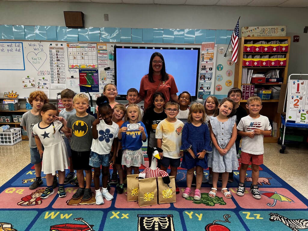 A smiling teacher stands in front of a classroom with a group of young students posing together on a colorful alphabet rug, with an American flag, posters, and classroom supplies in the background.