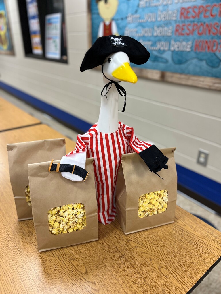 A a white plastic porch goose with a black pirate hat and yellow beak, wearing a red-and-white striped shirt, standing behind three brown paper bags filled with popcorn on a wooden table.