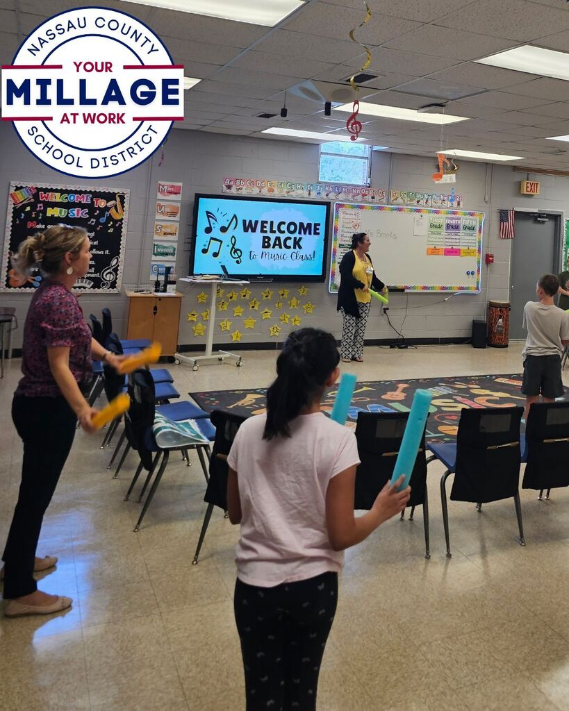 Students participate in a music class warm-up activity while standing and moving around the room with colorful rhythm sticks. The teacher leads the lesson from the front of the classroom near a display that reads “Welcome Back to Music Class!” The warm-up combines singing, vocal exercises, and coordinated movement to engage students in music learning.