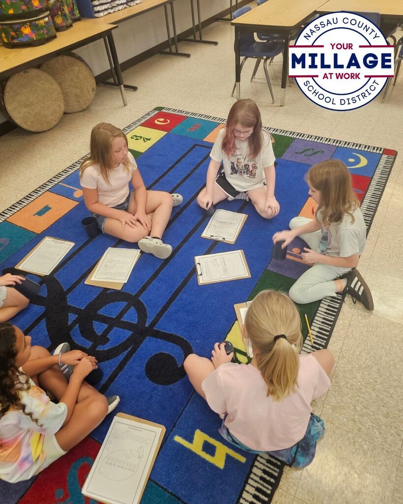 A small group of elementary students sits in a circle on a colorful music rug, using cups and worksheets to practice rhythm exercises. The activity encourages students to follow musical patterns and work together to develop timing and coordination skills.