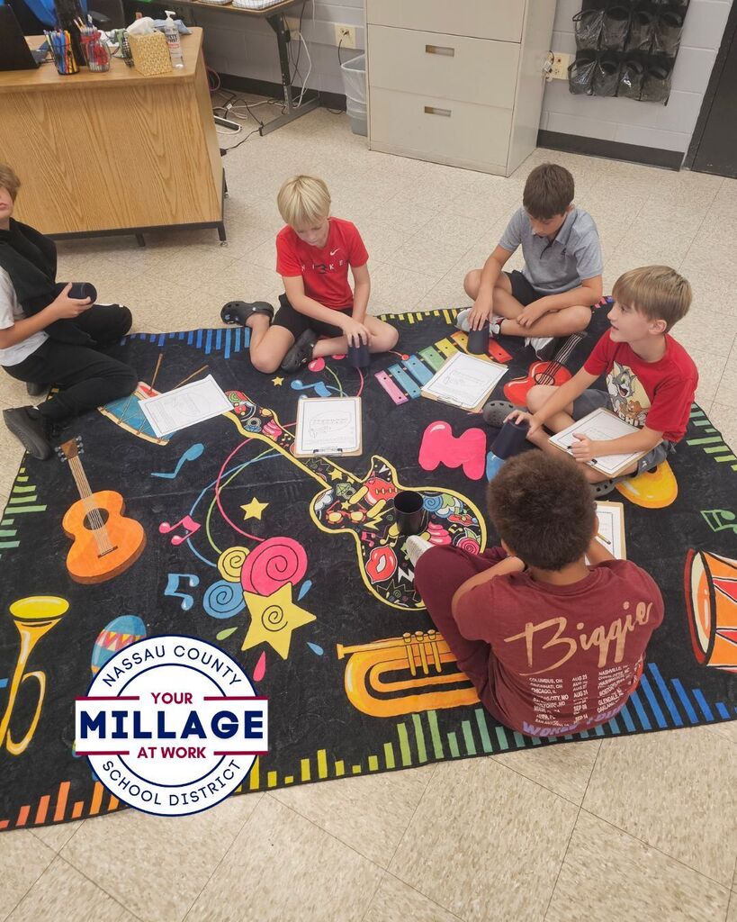 A small group of elementary students sits in a circle on a colorful music rug, using cups and worksheets to practice rhythm exercises. The activity encourages students to follow musical patterns and work together to develop timing and coordination skills.
