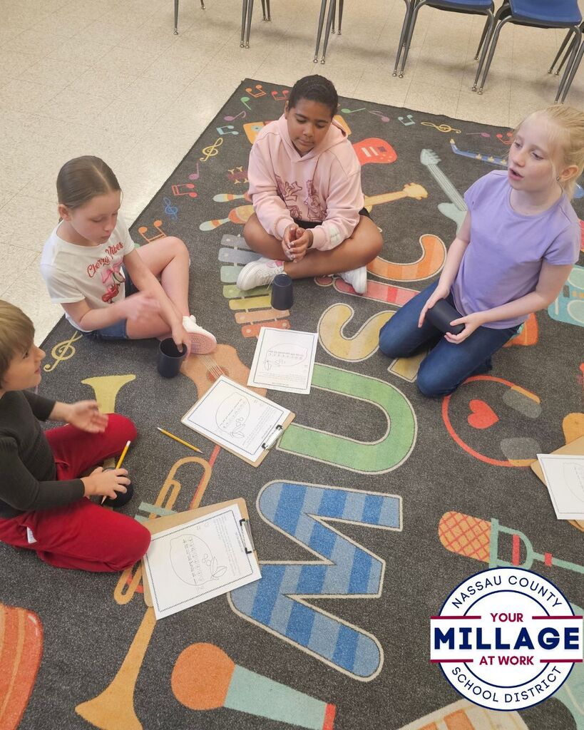 Four elementary students sit on a music-themed classroom rug participating in a cooperative cup-rhythm activity. Students follow music assignments on clipboards while practicing rhythmic patterns with plastic cups.