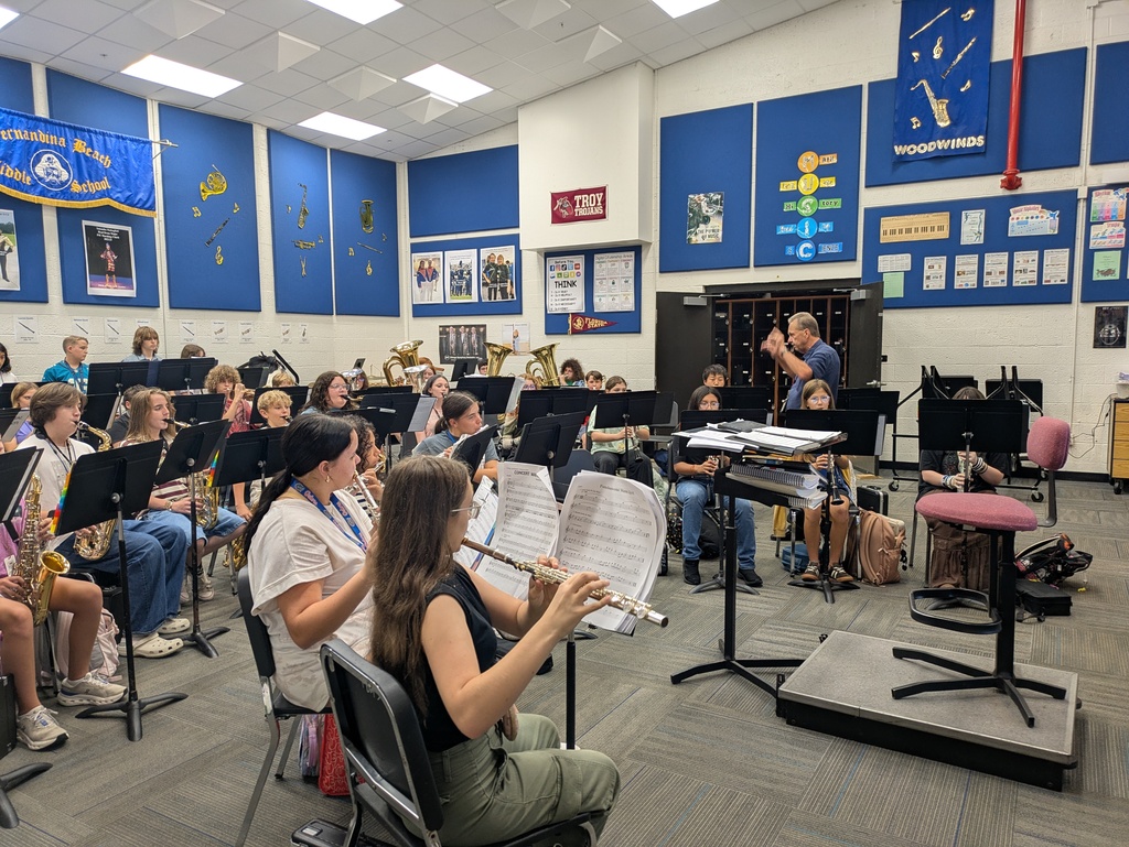 School band students playing instruments during rehearsal while the conductor leads the ensemble from the front of the room.