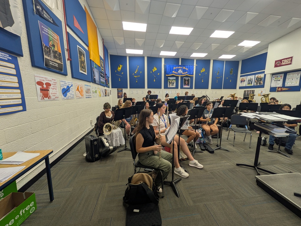 Students seated with instruments in a school band room during a music rehearsal, surrounded by music stands and classroom displays.
