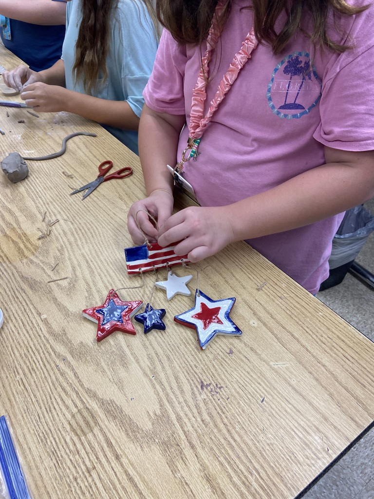 Student assembling red, white, and blue clay ornaments shaped like stars and an American flag at a classroom worktable.