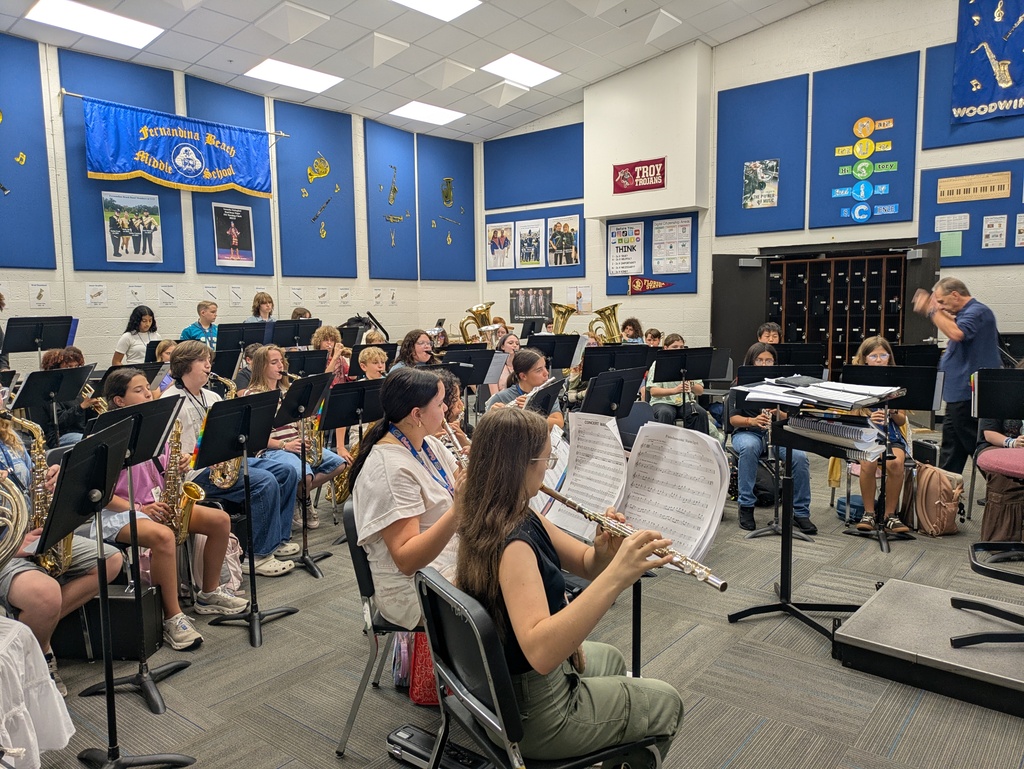 Students rehearse in a middle school band room while a conductor leads the ensemble.