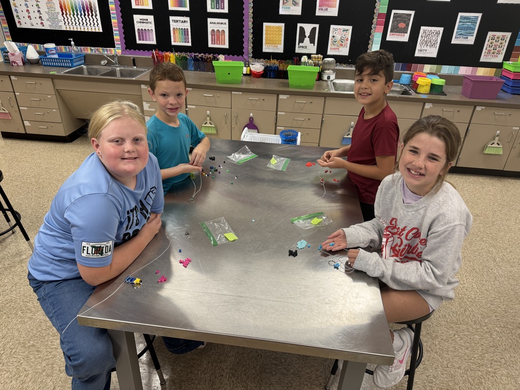 Four fourth-grade students work together at a table in the art classroom, using colorful beads and string to create handmade beaded keychains.