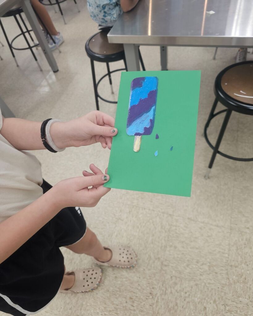 Student holds a colorful oil pastel artwork of a popsicle mounted on green construction paper in an art classroom. Additional images show student artwork displayed on a hallway wall and students using oil pastels at a classroom table while practicing blending and coloring techniques.