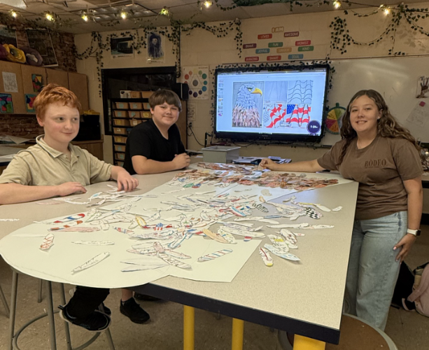 Three students sit and stand around a classroom table sorting and arranging decorated paper feathers. A digital display behind them shows artwork featuring a bald eagle and an American flag, which appears to serve as inspiration for the collaborative project.