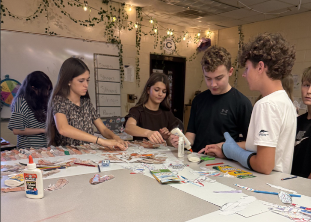 Several students collaborate around an art classroom table covered with paper feather cutouts, markers, crayons, and glue. Students sort, decorate, and assemble the feathers as part of a large group art project.
