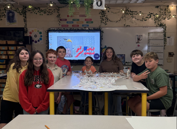 A group of students gathers around a classroom table displaying numerous decorated paper feathers assembled into a large eagle-shaped artwork. A digital display in the background shows a bald eagle and American flag design that corresponds with the project materials on the table.