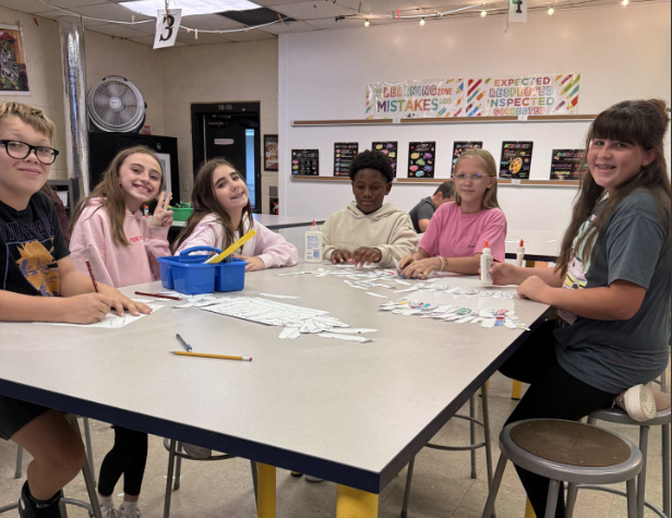 A group of students sits around a classroom table covered with paper feathers and art materials while contributing to a collaborative art project. Classroom artwork and posters are displayed on the walls in the background.