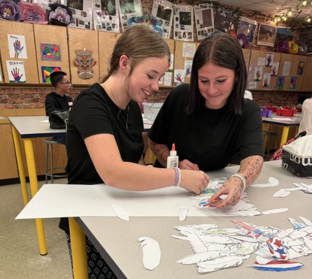Two students work together at an art classroom table, arranging and gluing decorated paper feathers onto a large display board. Additional colored feather cutouts and art supplies are spread across the table.