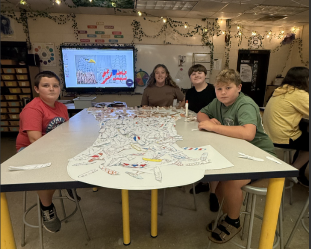 Students work together around a classroom table covered with paper feather cutouts for a large art project. A digital display in the background shows a reference image of a bald eagle and an American flag design that appears to be guiding the project.