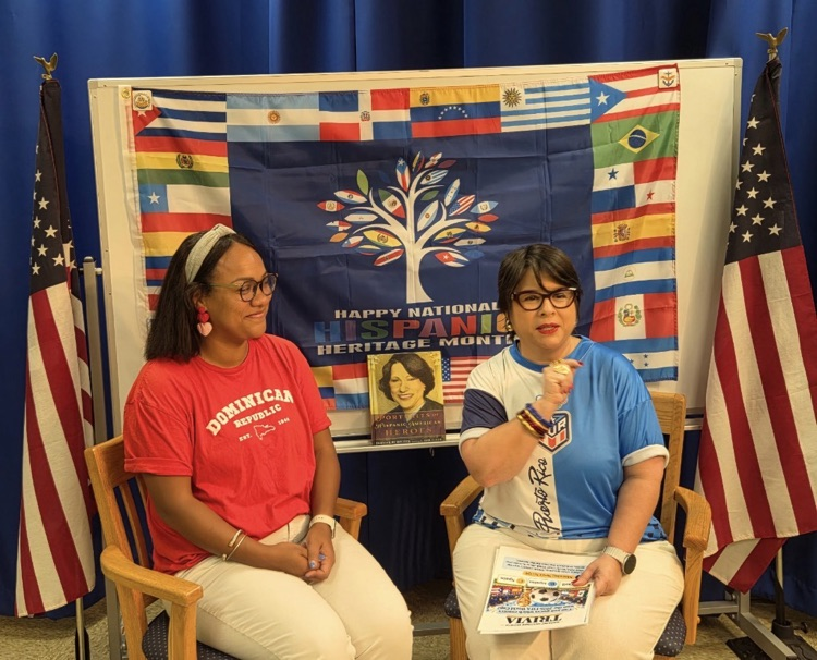 two Hispanic women are sitting in front of a flag displaying various Hispanic countries.  there are also two large American flags in stands on each side of them.  they are wearing Dominican and Puerto Rico shirts