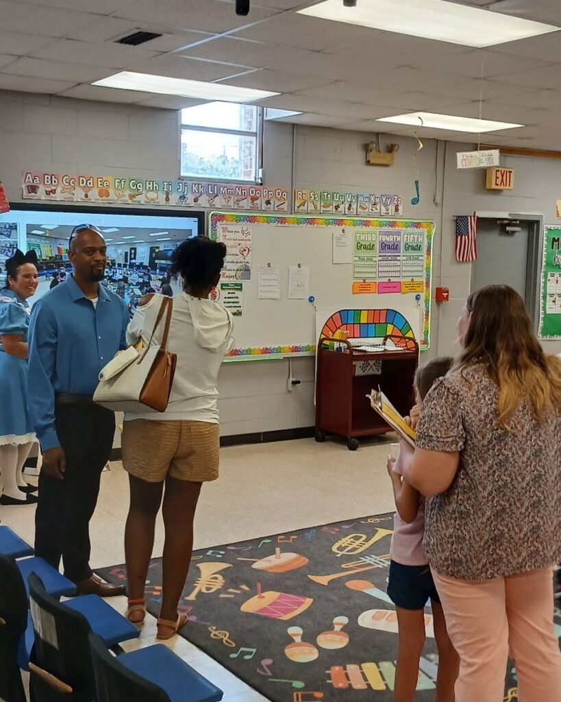 image shows families exploring the music room while a teacher dressed as Alice in Wonderland welcomes guests and shares examples of student learning.