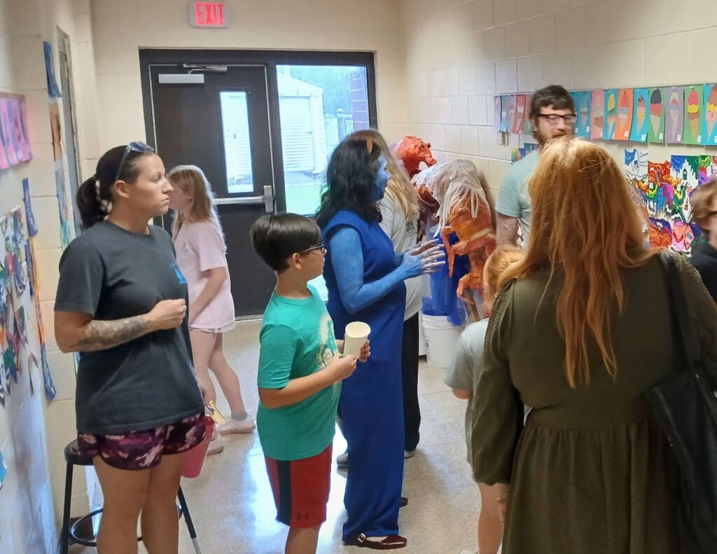 image shows families viewing colorful student artwork in the art hallway while a teacher dressed as Violet Beauregarde from Willy Wonka engages visitors and discusses student art projects
