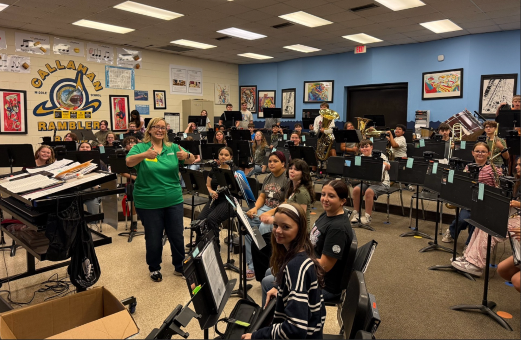 Students sit in a middle school band room during rehearsal, surrounded by music stands and instruments including flutes, clarinets, brass instruments, and percussion. An instructor stands near the front of the room, while students are seated in rows facing forward. The walls display student artwork, music-related posters, and a large “Callahan Ramblers” school logo. Bright overhead lighting illuminates the classroom.