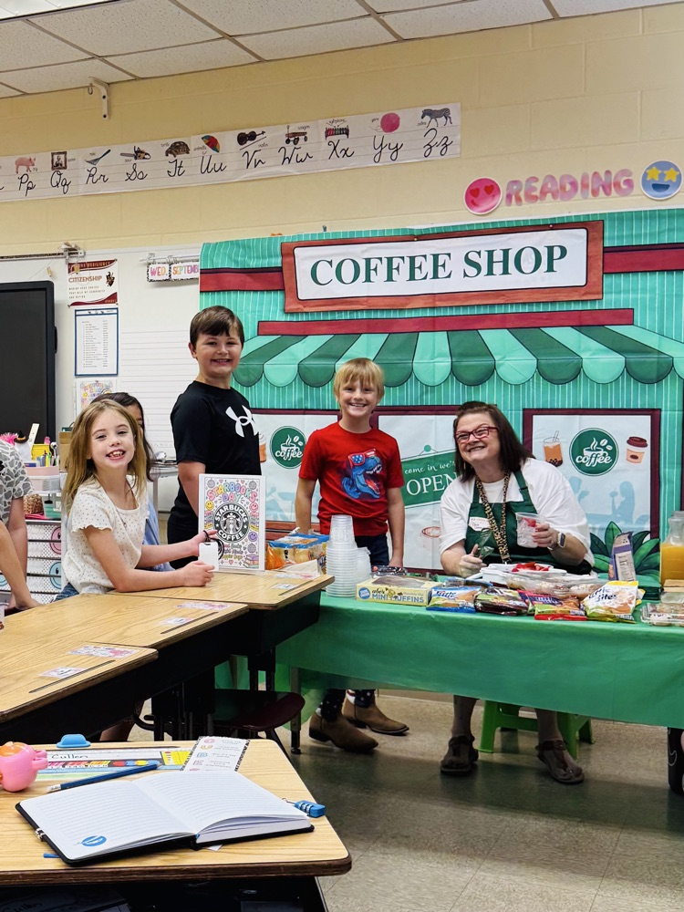 Students smile alongside their teacher during a fun Starbooks classroom experience. A coffee-shop-themed setup with books and treats helps make reading extra special.
