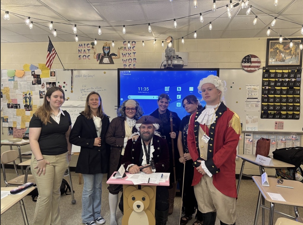 Seven students and a teacher in Mr. Bryan’s classroom pose in colonial-era costumes while celebrating Constitution Day. Several are dressed as historical figures from the Constitutional Convention, with patriotic classroom decorations and an American flag visible in the background.