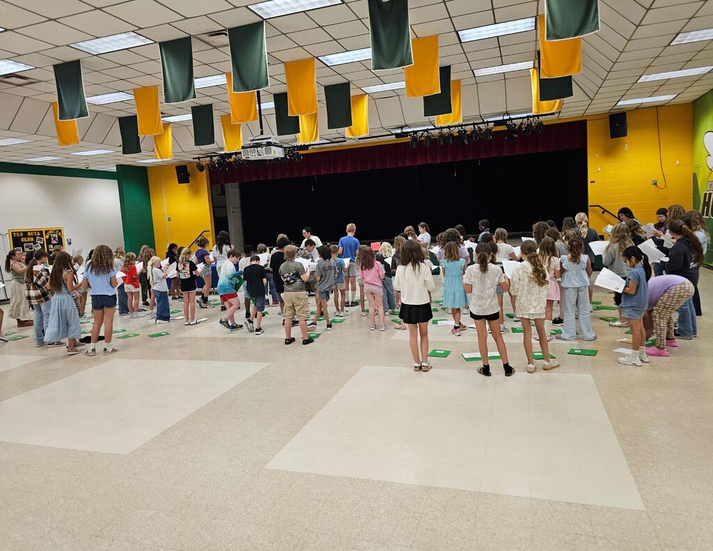 Students stand in rows in the Yulee Elementary cafeteria rehearsing for Hornet Singers, the school's after-school chorus program. Students hold music sheets while practicing vocal techniques and performance skills under the guidance of instructors. A stage with curtains is visible in the background, and green and gold banners hang from the ceiling.