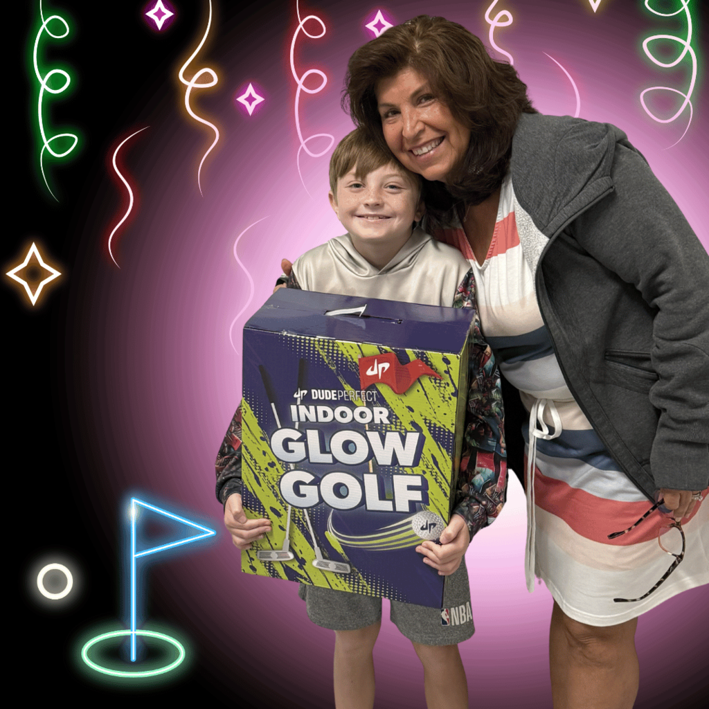 A 4th grade boy is posing with his teacher.  He is holding a large box title Indoor Glow Golf.  The picture includes decorative images of glow  golf images and glow decorative images making the photo background  celebratory. 