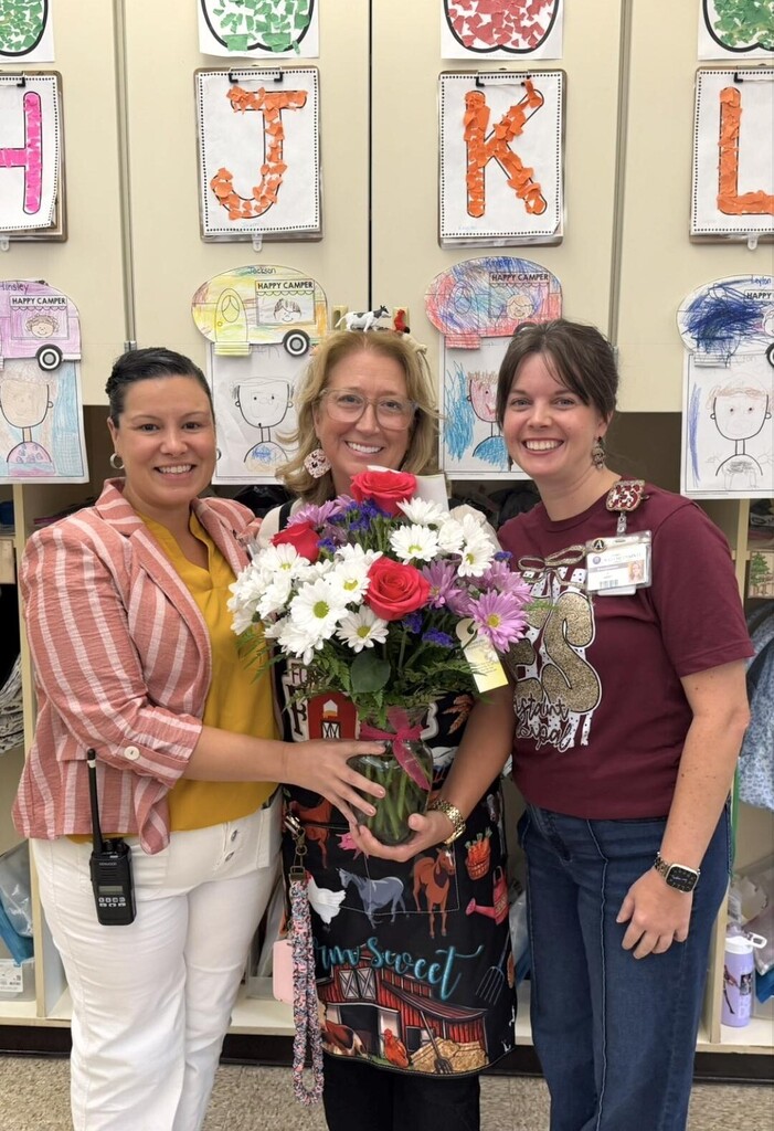 Three smiling women stand in a classroom, holding a vibrant floral arrangement with red roses and white daisies.
