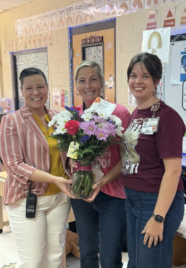 Three smiling women stand in a classroom, holding a vibrant floral arrangement with red roses and white daisies.