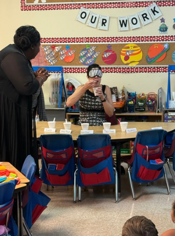 A teacher stands beside a classroom table while a presenter in a patterned dress gestures with both hands; children sit in blue chairs facing them, with planets on the wall and “OUR WORK” bunting above.