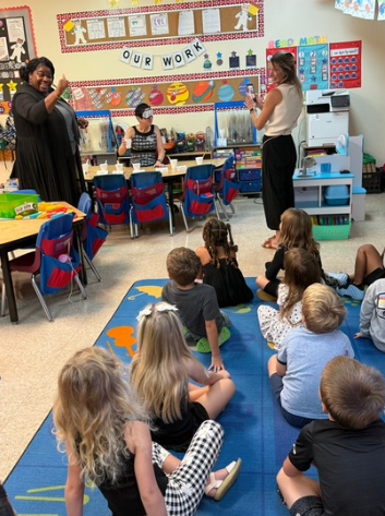A teacher stands beside a classroom table while a presenter in a patterned dress gestures with both hands; children sit in blue chairs facing them, with planets on the wall and “OUR WORK” bunting above.