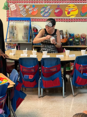 A person wearing a striped blindfold stands behind a table in a shop, holding a spoon, while blue-and-red chair covers line the front and colorful wall displays hang behind.