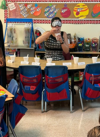 A person wearing a striped blindfold stands behind a table in a shop, holding a spoon, while blue-and-red chair covers line the front and colorful wall displays hang behind.