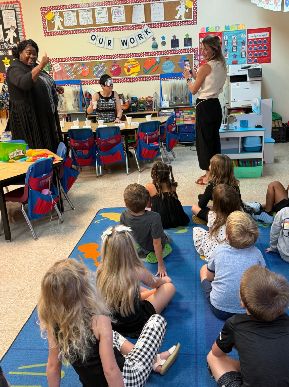 In a classroom, a teacher and an adult stand at the front while a group of children sit on a blue rug facing them; colorful posters and bins line the walls behind.