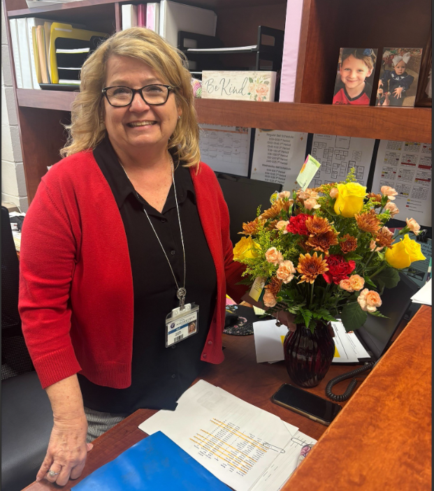 An individual stands behind a desk in an office workspace next to a large floral arrangement in a dark glass vase. The bouquet contains yellow roses, red flowers, peach-colored blooms, and bronze chrysanthemums with green foliage. Papers, a phone, a computer monitor, shelves with binders, and framed photographs are visible in the office background.