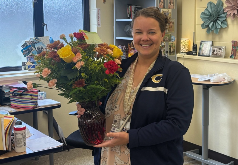 A person stands in an office classroom holding a large bouquet of colorful flowers in a dark glass vase. The bouquet includes yellow roses, pink flowers, red blooms, and autumn-colored chrysanthemums. Desks, shelves, framed photos, and paper decorations are visible in the background."