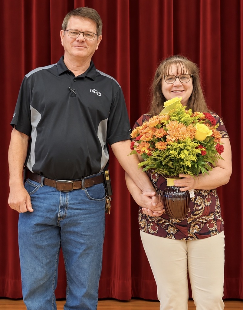 A man and woman stand together on a wooden stage in front of a closed red theater curtain. The woman smiles while holding a large arrangement of yellow, orange, and red flowers. The pair are holding hands and posing for a recognition photo.