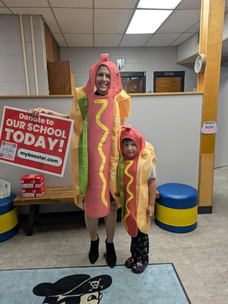 Two people in hot-dog Halloween costumes stand indoors, smiling on a pirate-themed rug; one holds a red “Donate to OUR SCHOOL TODAY!” sign while the other wears the same costume and a child stands beside them.