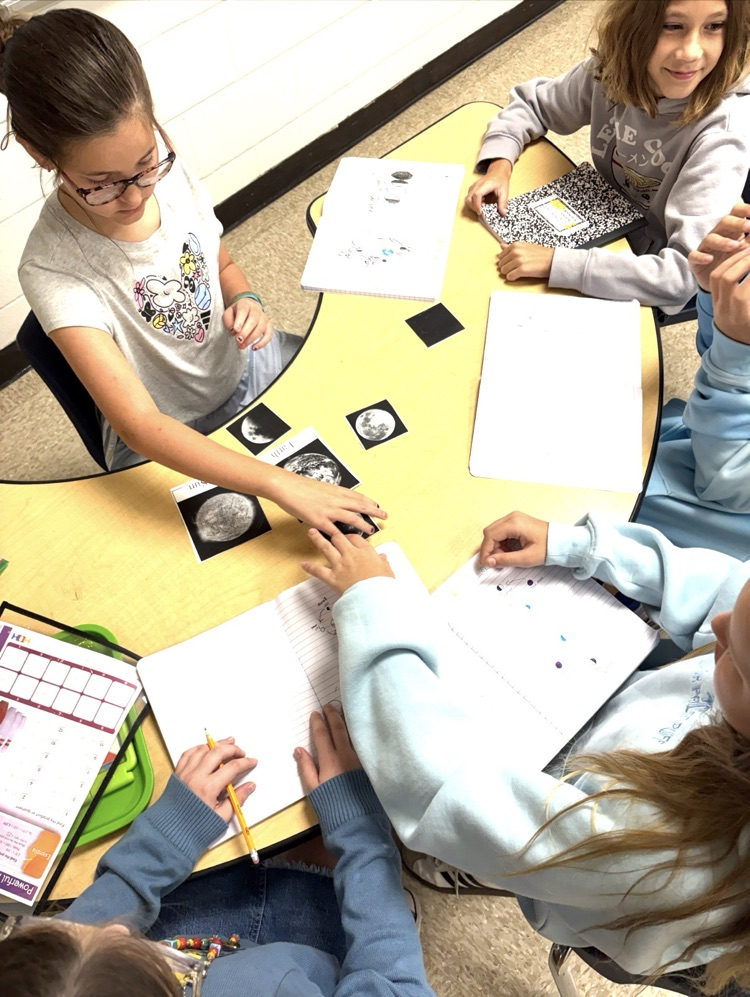 students sitting at a table with journals and pencils.  students are working together with phases of the moon cards