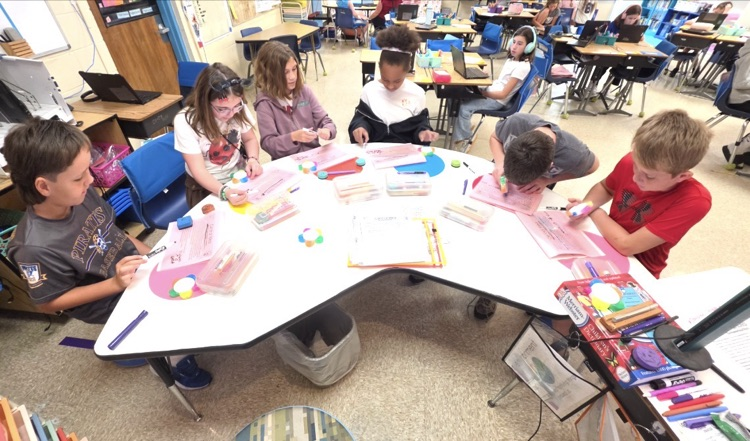 a group of students are sitting at a kidney shape table utilizing tools such as dry erase boards and highlighters while working on comprehension 