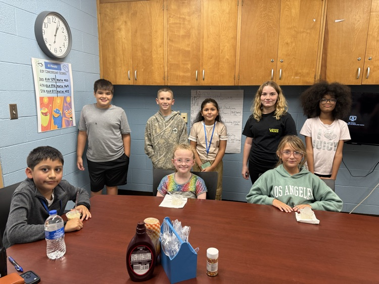 a group of students smiling for the camera.  2 students are sitting at a large conference table enjoying ice-cream.  4 additional students are standing 
