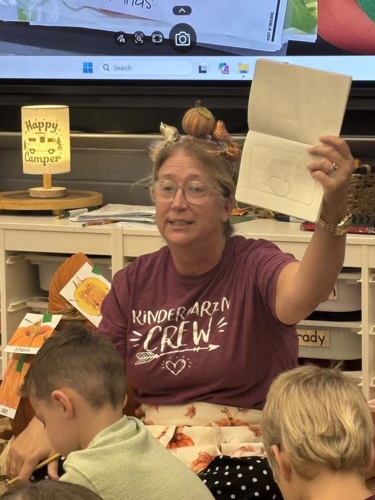 Kindergarten teacher sits with students and holds up a student paper during a pumpkin science lesson. Small decorative pumpkins are tucked into her hair as part of the classroom activity.