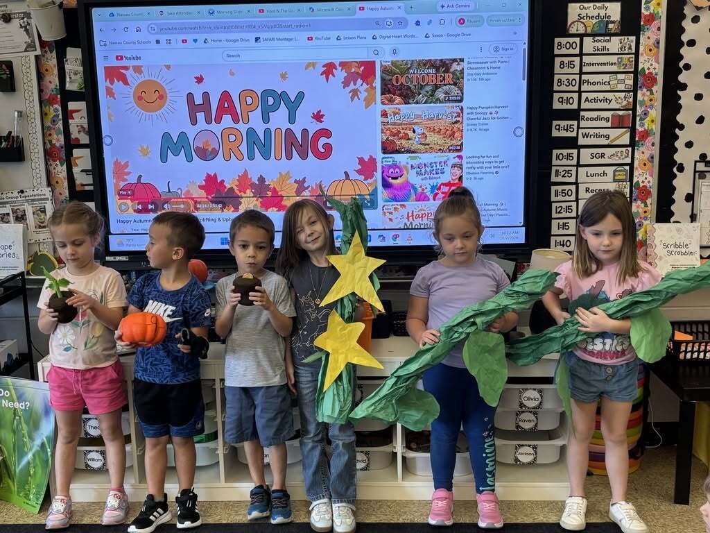 Six Callahan Elementary kindergarten students stand at the front of their classroom holding props representing stages of the pumpkin life cycle, including a sprout, pumpkin, vine, and flower.