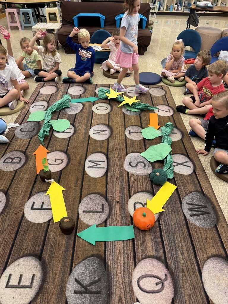 Kindergarten students sit around a large floor model of the pumpkin life cycle made with pumpkins, vines, flowers, leaves, and arrows showing the sequence of growth.