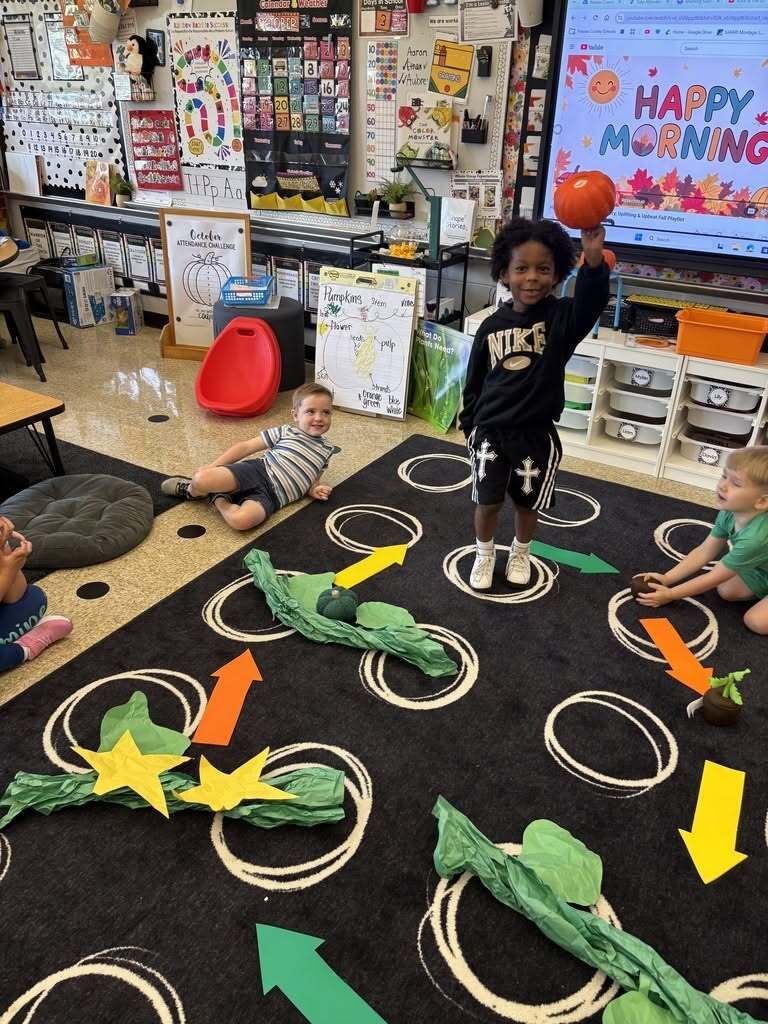 Kindergarten student holds a pumpkin above his head while classmates sit around a large floor model illustrating the stages of the pumpkin life cycle.