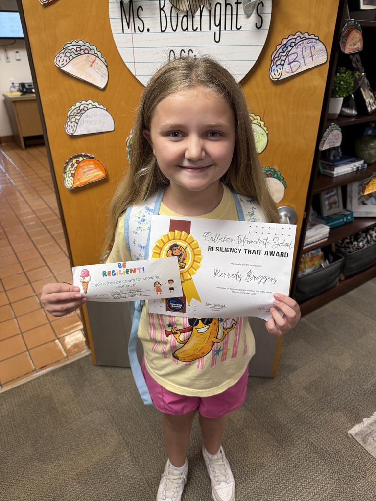 A smiling elementary school student stands in front of a classroom door, holding a Resiliency Trait Award certificate recognizing her for demonstrating the trait of honesty, along with an ice cream reward certificate for showing resilience.