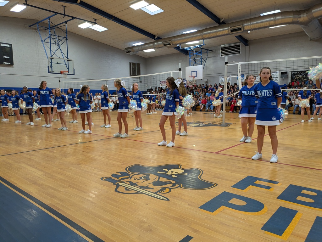 Cheerleaders in blue uniforms stand in formation on a gymnasium floor during a school event.