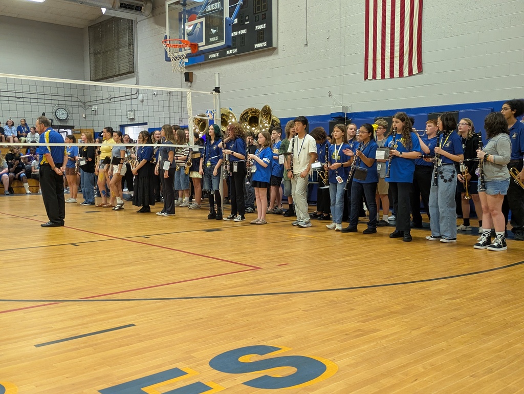 School band members stand with instruments in a gymnasium during a school assembly.